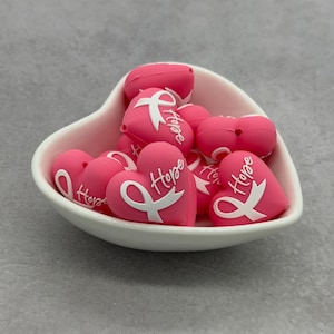 May include: A white heart-shaped bowl filled with multiple pink heart-shaped beads. Each bead features a raised white awareness ribbon and the word Hope embossed on the surface. The beads are smooth and matte, designed with a central hole for stringing.