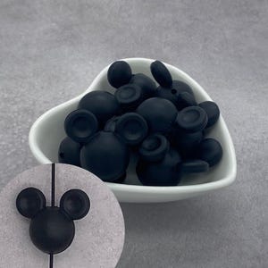 Puede incluir: Una colección de cuentas negro mate con forma de cabezas de ratón con dos orejas circulares y una cabeza redonda central. Las cuentas están contenidas en un cuenco blanco en forma de corazón, con una cuenta mostrada en detalle en un recuadro.