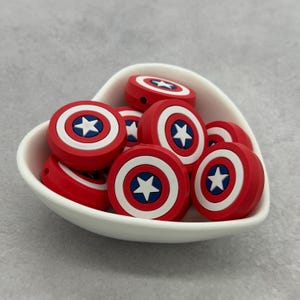 May include: A collection of round, red, white, and blue shield-shaped beads, each featuring a central white star on a blue circular background, contained within a white heart-shaped bowl. The beads have a thick, cylindrical profile.