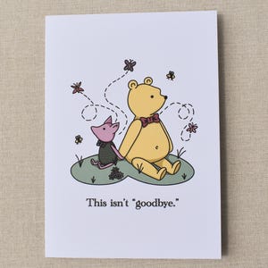 May include: A greeting card featuring an illustration of a yellow bear and a pink pig sitting together on a patch of green grass. Butterflies and flowers are drawn around them. The text "This isn't "goodbye."" is printed at the bottom.