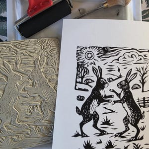 May include: A black and white linocut print depicting two hares dancing, with a carved linoleum block, a red handled brayer, and a carving tool. The hares are set against a backdrop of trees, sun, and clouds. The print is on white paper.