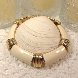 May include: Cream-colored enamel bracelet with gold-tone accents. The bracelet is a rounded rectangular shape with gold bands at intervals. The bracelet is resting on a large, white seashell.