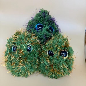 May include: Three plush toys shaped like balls with green and gold fur and large blue eyes. The top toy has purple and green fur.
