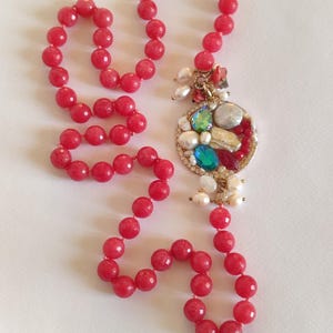 Può includere: Una collana lunga con perline sfaccettate rosa brillante, che porta a un ciondolo circolare color oro. Il ciondolo è decorato con un gruppo di perle bianche, piccole perline rosse e pietre sfaccettate dai colori vivaci blu e verde.