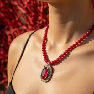 May include: A red beaded necklace with a large oval pendant. The pendant features a large, red gemstone surrounded by a decorative frame with small, clear stones. The necklace is worn against a black top.