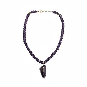 Puede incluir: Un collar de cuentas moradas con un colgante. El collar presenta pequeñas cuentas facetadas en un tono morado intenso. Un colgante más grande, de forma irregular, cuelga del centro, mostrando una mezcla de tonos morados y marrones. Tiene un cierre dorado.
