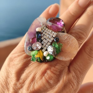 Può includere: Un anello grande e ornato con un gruppo di pietre preziose colorate, perle e strass. L'anello ha una base a forma di cuore con una grande gemma viola e un accento a forma di diamante argentato. L'anello è su una mano.