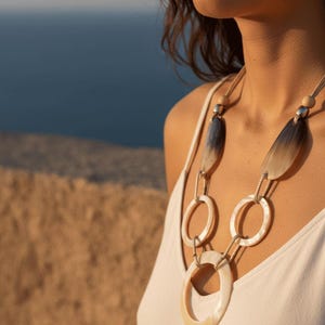 Peut inclure: Un collier de déclaration avec de grands pendentifs ovales et circulaires aux tons crème et gris. Le collier est suspendu à un cordon beige et à des maillons en métal argenté. Le modèle porte un débardeur blanc.