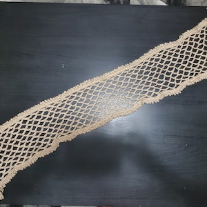 May include: A beige crocheted table runner with fringe on the ends. The runner is made of a delicate, open-weave pattern.