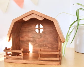 May include: A wooden barn playset with a small window and a fence around the front. The barn is made of light brown wood and has a natural wood finish.