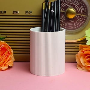 May include: A white cylindrical pencil holder with a vertical ribbed texture, containing several black pens. The holder is positioned in front of a vintage olive-green Bush radio and flanked by two orange artificial flowers on a pink surface.