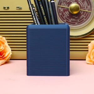 May include: A navy blue pencil holder with a ribbed texture, filled with pencils, sits in front of a vintage radio. The radio is beige with a gold-coloured dial and a pink background. Orange roses are on either side.