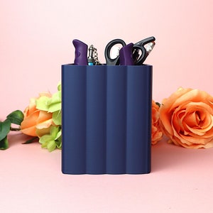 May include: A dark blue desk organiser with four compartments, holding various office supplies, including scissors, pens, and a key chain. The organiser is sitting on a pink surface.