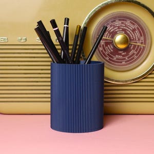 May include: A dark blue pencil pot with vertical grooves, containing black pencils and a few gold and pink ones. The pot is on a pink surface, with a vintage radio in the background.