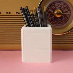 May include: A white square pencil holder with a horizontal ribbed texture containing several black pencils, including one labelled Western Woods 2B, and a few pencils with metallic gold and silver ends, standing upright in a cluster.