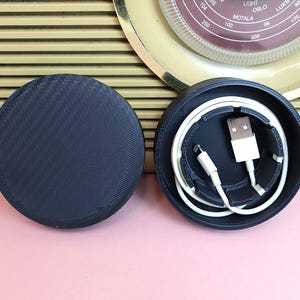 May include: Two black round cases, one with a white cable and a white USB connector inside. The cases are on a pink surface.