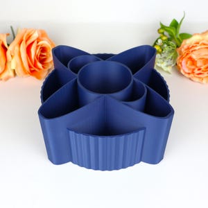 May include: A navy blue desk tidy with multiple compartments. The organiser has a central cylindrical section surrounded by petal-shaped compartments and a ribbed exterior. The product is made of plastic and is designed to hold pens, pencils, and other office supplies.