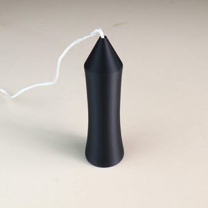 May include: A matte black, cylindrical light pull with a tapered, conical top section. The object features a smooth, slightly concave body and is attached to a thin, white braided cord that extends from the centre of the pointed top.
