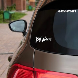May include: A white vinyl sticker with the text "KaiWachi" on a black car window. The sticker is on the back window of the car. The text is in a handwritten style.