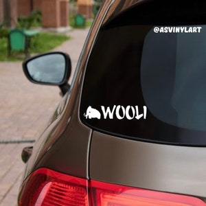 May include: A white vinyl sticker with a silhouette of a woolly mammoth and the word "WOOLI" in a stylized font. The sticker is applied to the back window of a brown car.