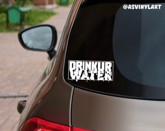 Può includere: Adesivo in vinile bianco con il testo "DRINK UR WATER" in un font a goccia, applicato sul lunotto posteriore di un'auto marrone. L'adesivo è su uno sfondo nero.