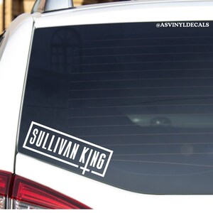 May include: White vinyl decal with the text "Sullivan King" in a rectangular frame with a white cross underneath. The decal is on the back window of a white car.