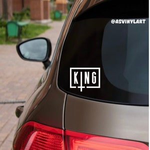 May include: A black car with a white vinyl sticker on the back window that says "KING" in a box with a cross underneath.