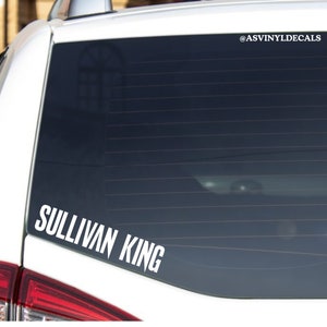 May include: White vinyl decal with the text "Sullivan King" on a black car window. The decal is placed on the lower right corner of the window. The text is in a bold, sans-serif font.