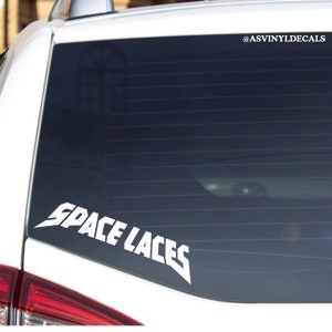May include: White vinyl decal with the text "SPACE LACES" in a bold, stylized font. The decal is applied to the rear window of a white car. The decal is a popular design for car enthusiasts.