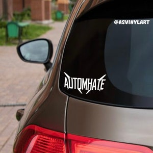 May include: A black car with a white vinyl sticker on the back window that says "AUTOMHATE" with a lightning bolt design. The sticker is on the lower right side of the window. The car is parked outside on a sunny day.