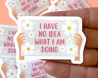 May include: A set of stickers with a white background and pink text that reads "I have no idea what I am doing". The stickers feature a daisy design and two hands holding the sign.