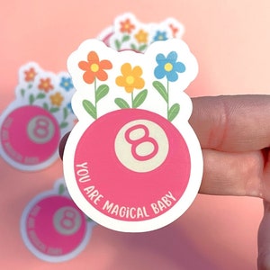May include: A pink 8-ball with a white number 8 on it. The 8-ball is surrounded by flowers in shades of blue, yellow, and orange. The text "You are magical baby" is written around the 8-ball.