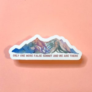 May include: A sticker with a watercolor illustration of a mountain range with the text "ONLY ONE MORE FALSE SUMMIT AND WE ARE THERE" in pink.