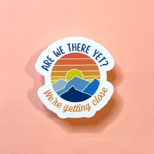 May include: White sticker with a sunset and mountains graphic. The text reads "Are we there yet? We're getting close."