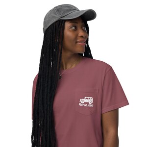 May include: A maroon t-shirt with a pocket displaying a white vehicle graphic and the text "NOMAD.ISHHH". The outfit includes a grey, faded baseball cap. A necklace is also visible.