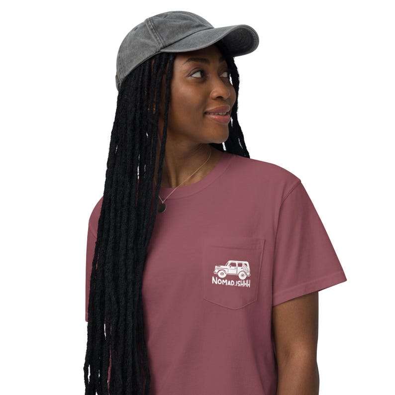 May include: A maroon t-shirt with a pocket displaying a white vehicle graphic and the text "NOMAD.ISHHH". The outfit includes a grey, faded baseball cap. A necklace is also visible.
