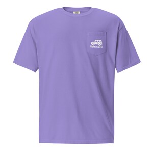 May include: A lilac t-shirt with a pocket on the left chest. The pocket displays a white graphic of a vehicle and the text "NOMAD ISH". The shirt has short sleeves and a crew neck.