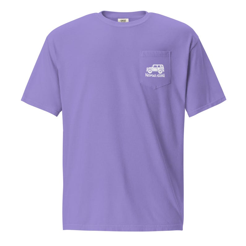 May include: A lilac t-shirt with a pocket on the left chest. The pocket displays a white graphic of a vehicle and the text "NOMAD ISH". The shirt has short sleeves and a crew neck.