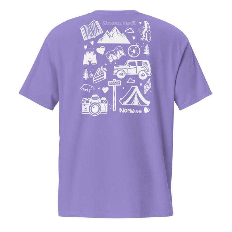 May include: A lilac t-shirt with a white graphic print of outdoor adventure icons, such as a tent, camera, and mountain range. The words "NATIONAL PARKS" and "NOMAD.ISH" are printed on the shirt.