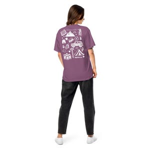 May include: A mauve t-shirt with a white graphic design on the back featuring mountains, a tent, a camera, and a jeep. The text "Nomad" is printed below the graphic. The person is wearing black jeans and white sneakers.
