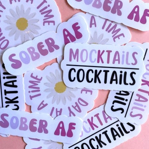 May include: A collection of stickers with various designs, including a daisy, the words "Sober AF", "Mocktails", and "Cocktails", and the phrase "A Time To Sober AF". The stickers are printed on white paper with pink and purple accents.