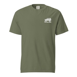 May include: Olive green short-sleeve t-shirt with a white graphic of a vehicle and the text "NOMADISHHH" on the upper right chest. The shirt has a crew neck and a relaxed fit.