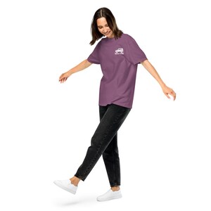 May include: A mauve t-shirt with a white graphic of a vehicle and the text "Never Quit" on the upper left chest. The person is wearing black jeans and white sneakers. The shirt is a solid color.