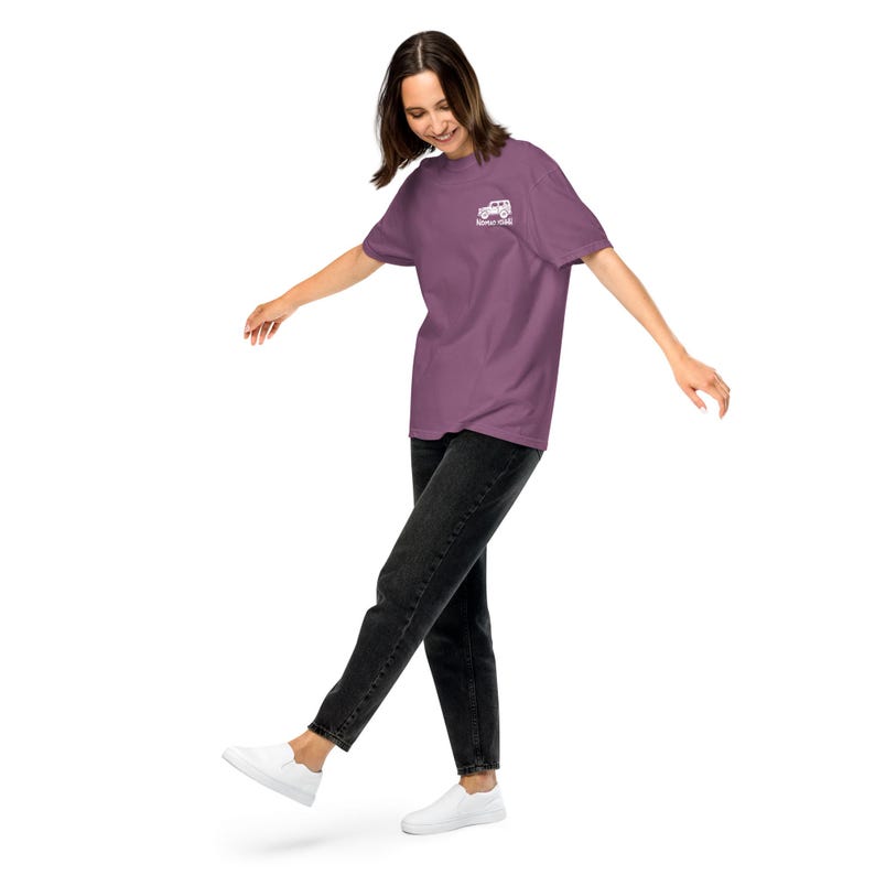 May include: A mauve t-shirt with a white graphic of a vehicle and the text "Never Quit" on the upper left chest. The person is wearing black jeans and white sneakers. The shirt is a solid color.
