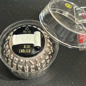 May include: A silver typewriter font ball with a black label that reads "Olde English" and a white label with a triangle and the text "10/12". The font ball is in a clear plastic container with a clear plastic lid.