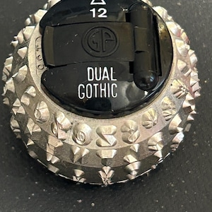 May include: A silver typewriter ball with black plastic casing. The ball has a triangular symbol with the number 12, a circular logo with the letters "GF", and the text "DUAL GOTHIC".