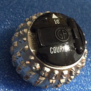 Puede incluir: Primer plano de un elemento de máquina de escribir plateado y negro. La esfera plateada tiene una superficie texturizada con formas geométricas en relieve. Una sección negra muestra el número "10" y la palabra "COURIER". Las letras "GP" también son visibles.