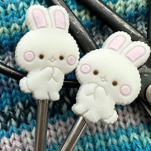 May include: Two silver knitting needles with white bunny-shaped ends. The bunnies have pink ears and brown eyes.