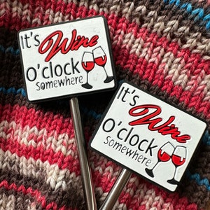 May include: Two silver wine bottle stoppers with white and red signs that say "It's Wine o'clock Somewhere" and feature a red wine glass graphic.