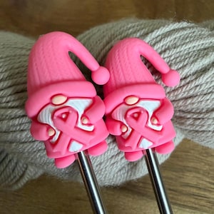 May include: Two identical crochet hooks featuring pink gnome toppers. Each gnome wears a textured pink hat and holds a pink breast cancer awareness ribbon against a white beard. The hooks have smooth, metallic silver shafts and rounded tips.